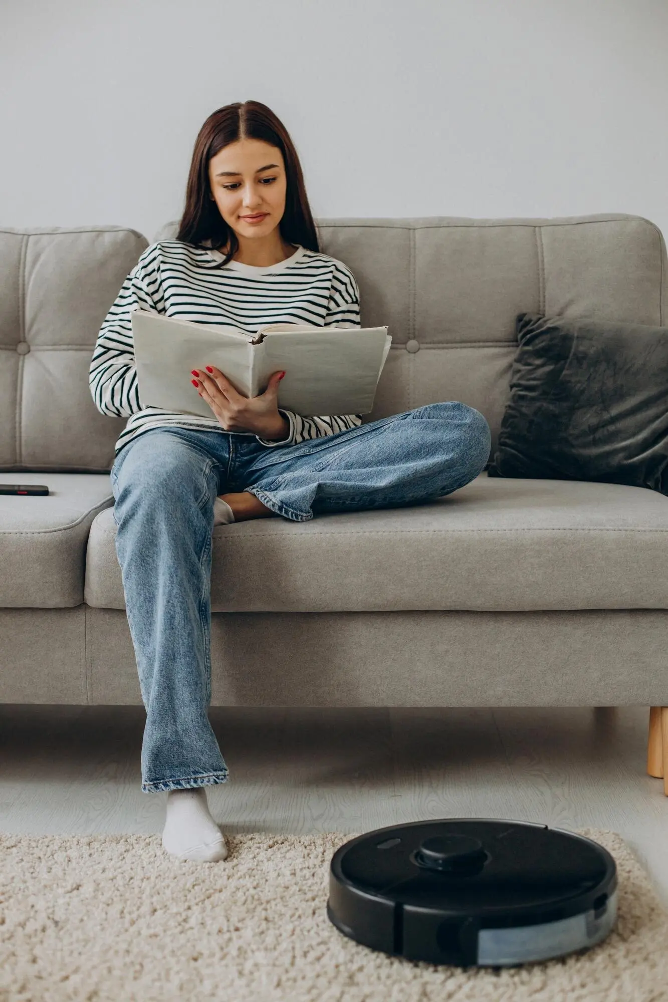 A woman sits on a sofa reading a book while a robot vacuum cleaner cleans the room.