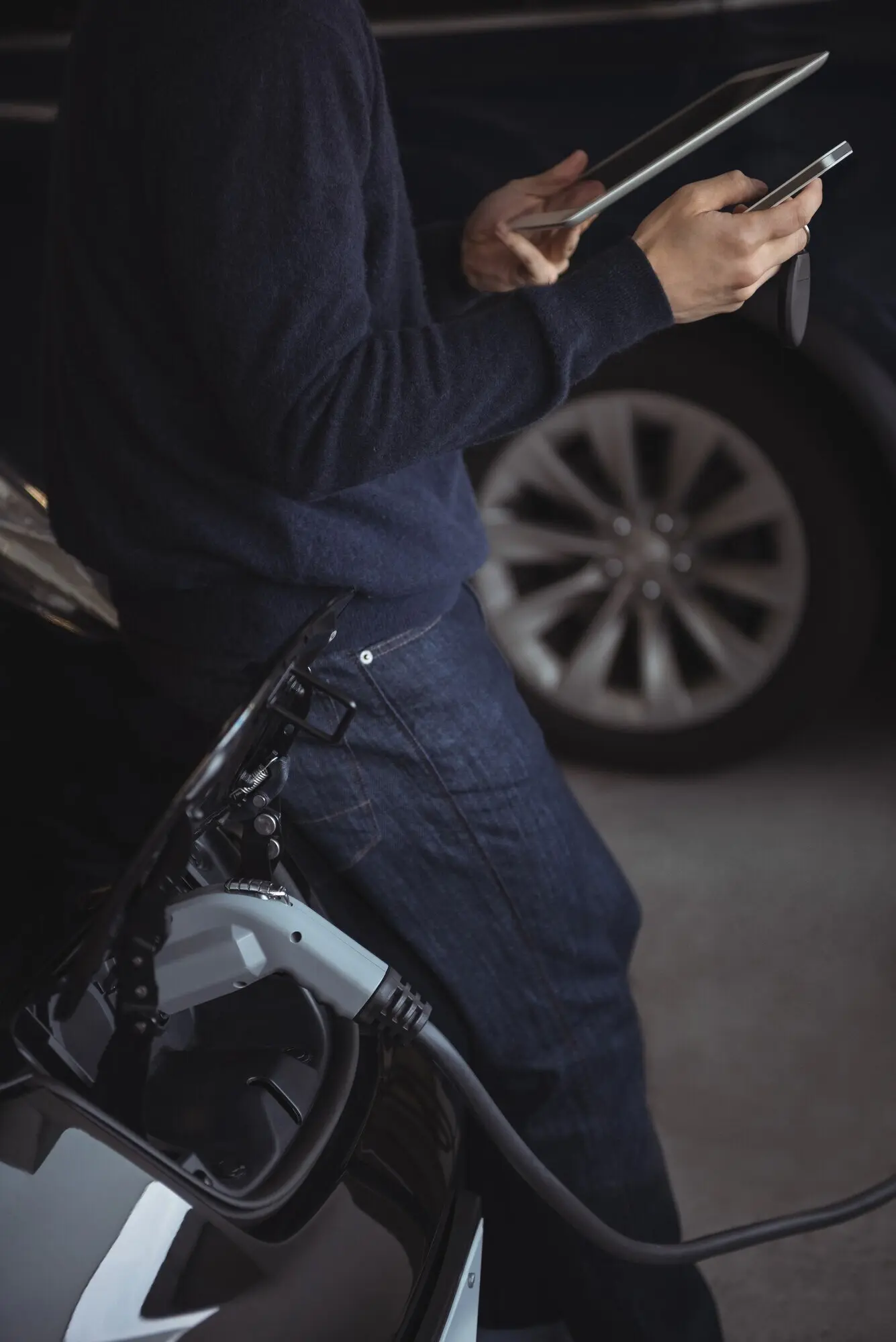 A man's midsection as he uses a digital tablet and a mobile phone while charging an electric car.