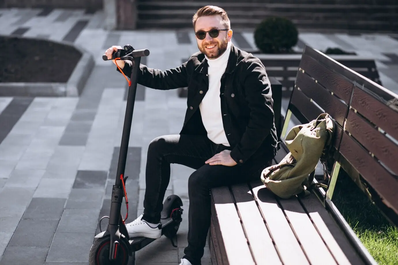 A handsome young man sits on a park bench, resting after a scooter ride.